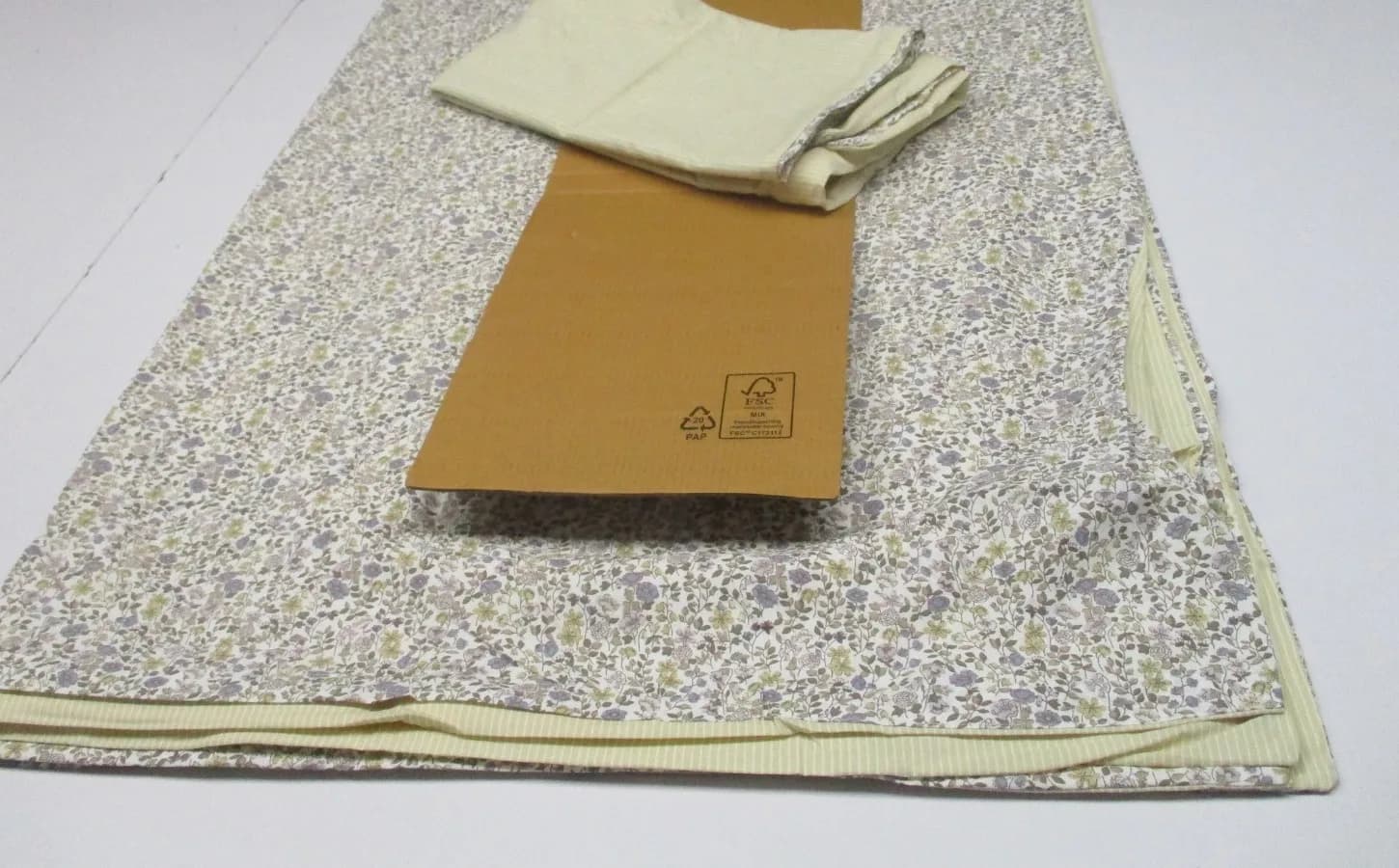 A folded floral cotton cover with a cream ribbed trim, laid flat on a white table beside a corrugated insert.