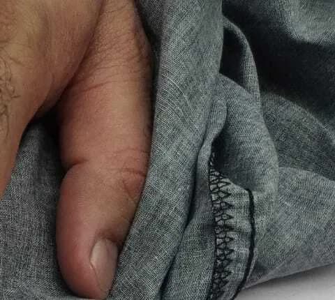 A hand gathering a length of grey woven cotton to test its drape, with the overlocked edge visible.