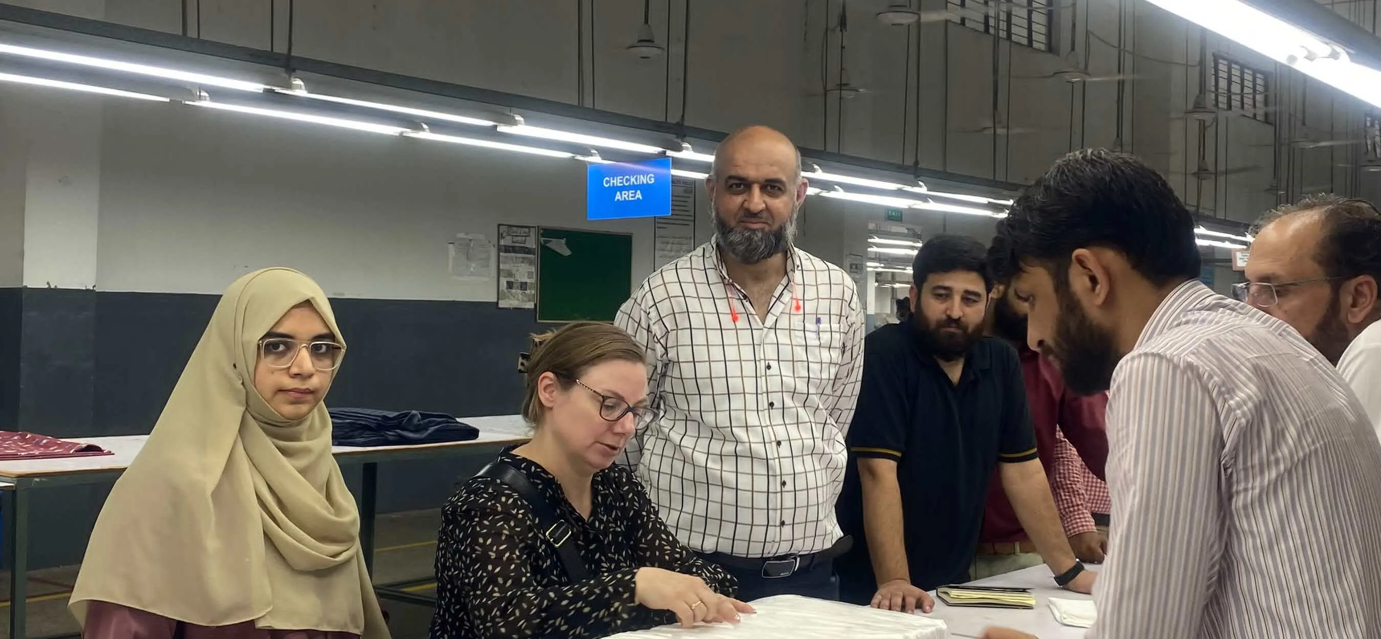 A group gathered around a long white inspection table on a factory floor, examining folded goods together.